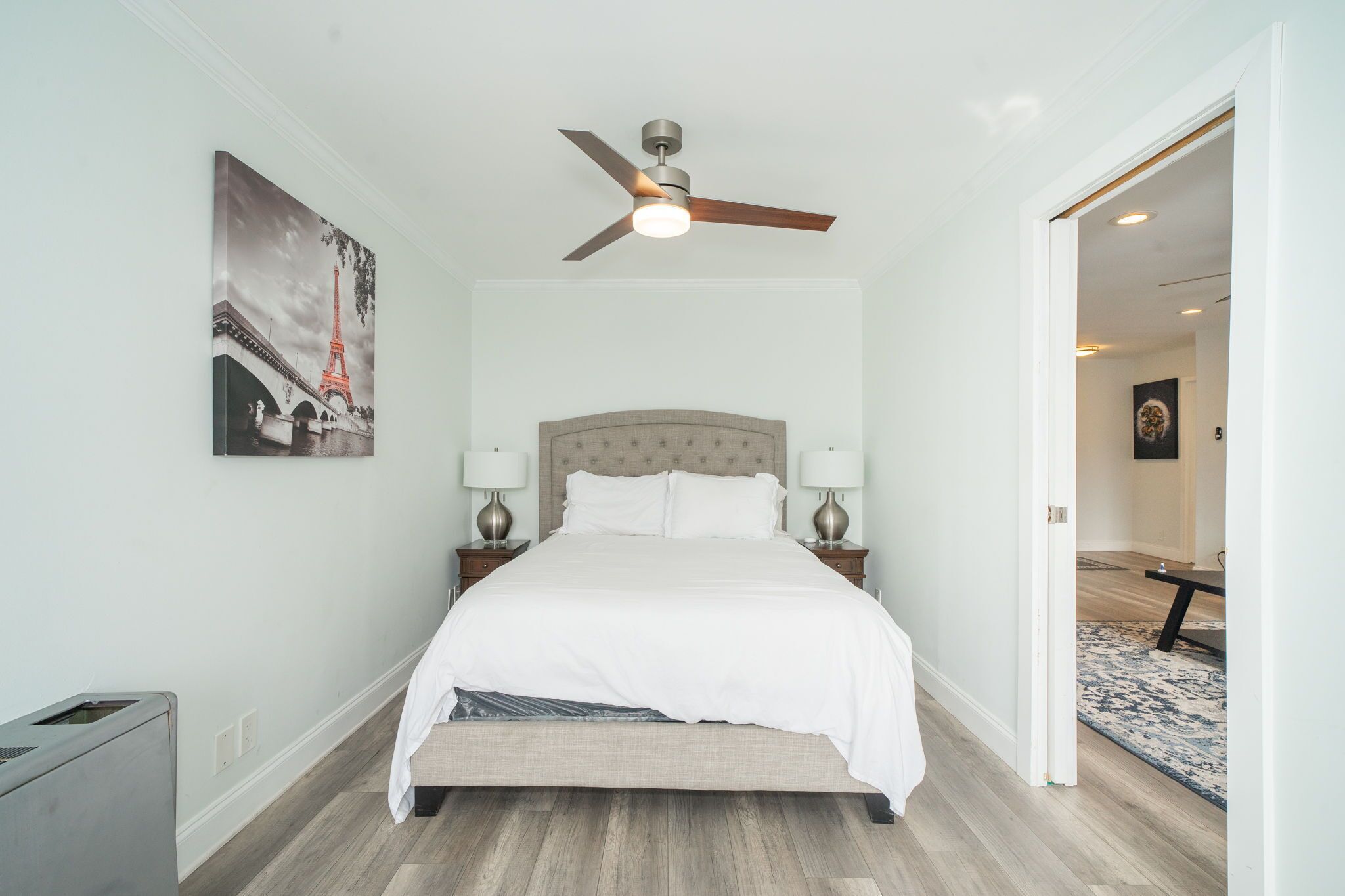 A bedroom with a bed, a ceiling fan, two bedside tables with lamps, and a painting of the Eiffel Tower.
