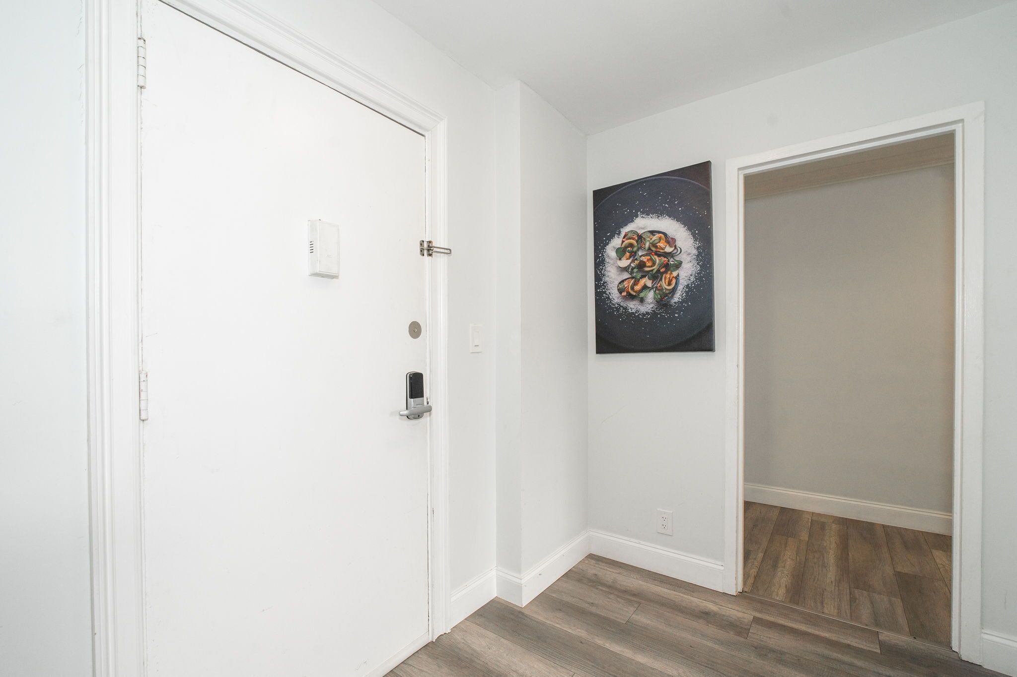A hallway with white walls, a wooden floor, a door with a lock, and a painting of a fruit bowl.