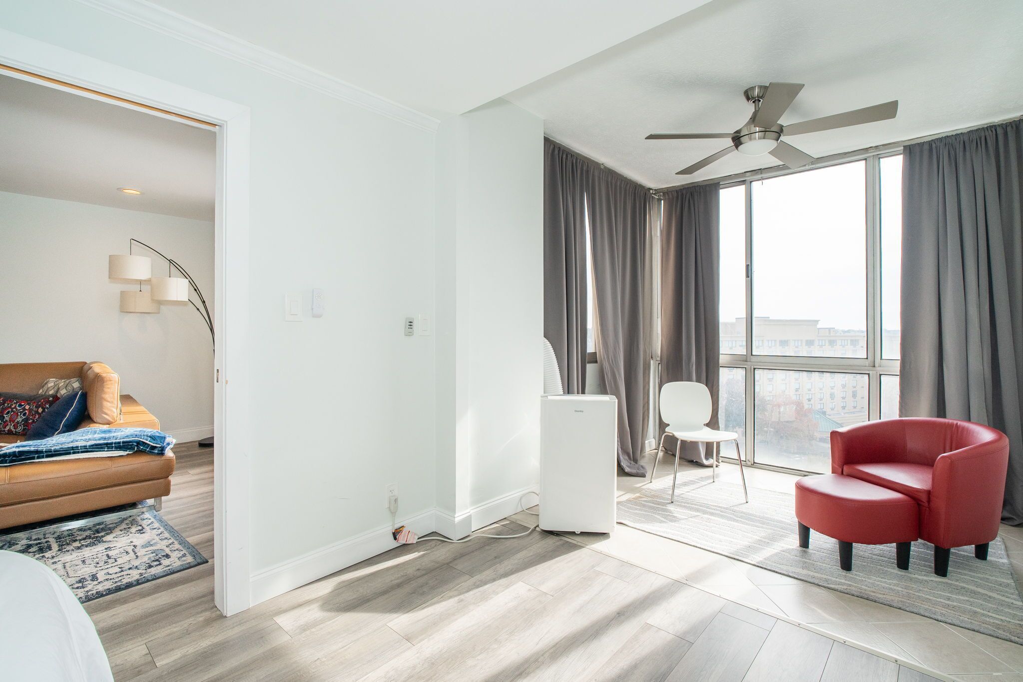 Une chambre d’hôtel moderne dotée d’une grande fenêtre, d’un ventilateur de plafond, d’un fauteuil rouge, d’un canapé blanc et d’un radiateur portatif.