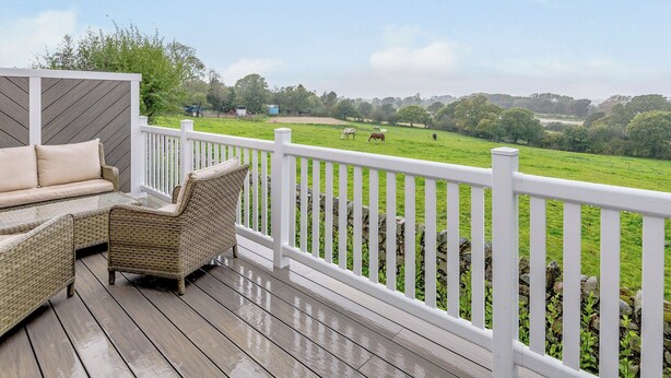 Eine Holzterrasse mit Korbmöbeln und einem weißen Geländer mit Blick auf eine grüne Wiese und Pferde.