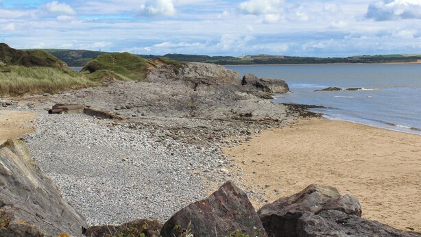 Paesaggio costiero con spiaggia di ciottoli, scogliere rocciose e colline in lontananza.
