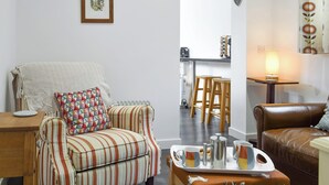 Un salon avec des meubles à rayures, une table basse sur laquelle se trouvent des tasses et une cafetière, et un coin cuisine à l’arrière-plan.