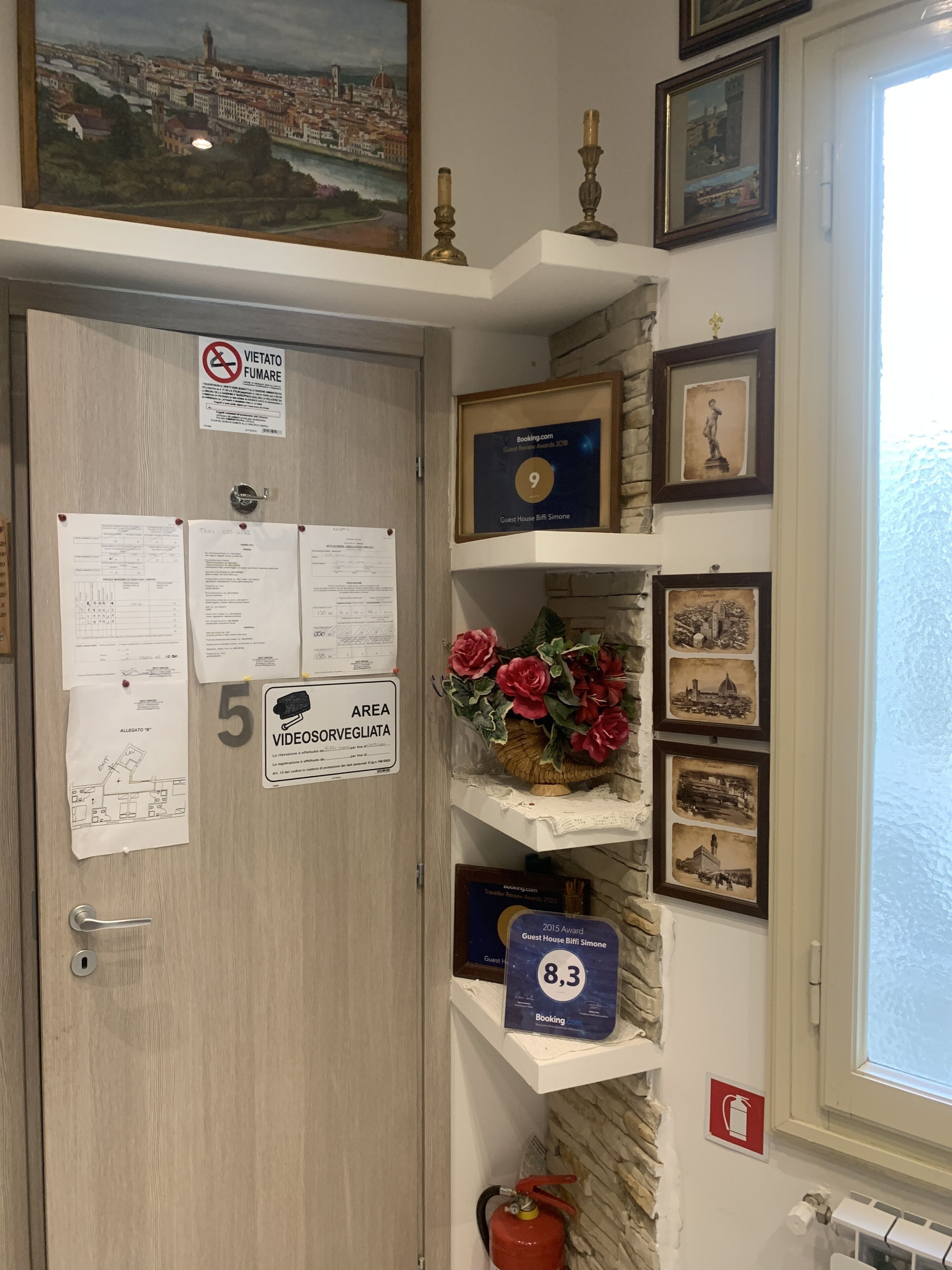 A hotel room with a wooden door, various notices, a framed certificate, a fire extinguisher, and a window with a view of a cityscape.