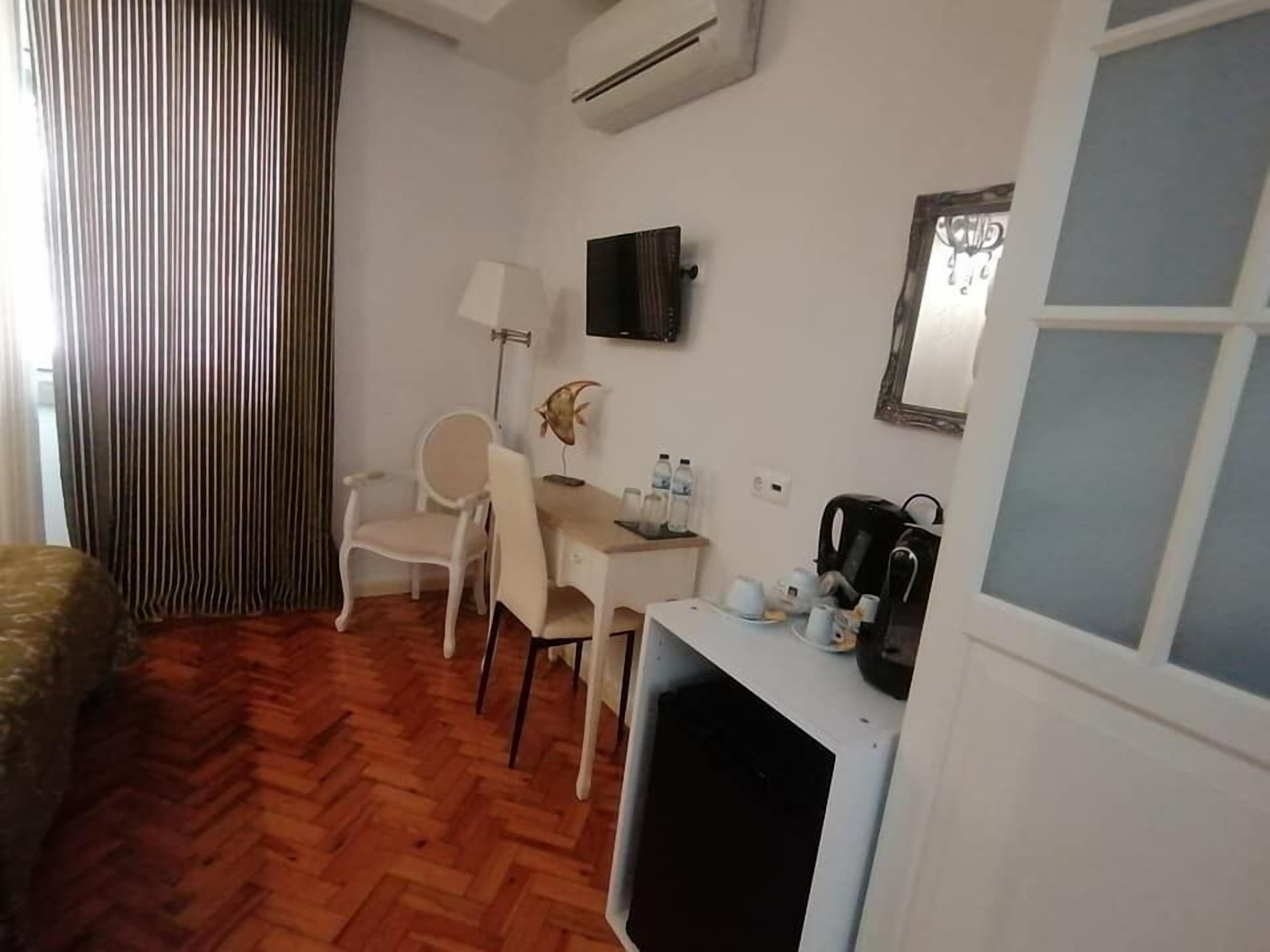 A hotel room with a wooden herringbone floor, a desk with a chair, a television mounted on the wall, a coffee machine, and a window with a view of the outside.