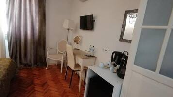 A hotel room with a wooden herringbone floor, a desk with a chair, a television mounted on the wall, a coffee machine, and a window with a view of the outside.