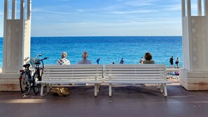 A covered outdoor seating area with white benches and a bicycle, overlooking a clear blue sea. - ✪ Stylish apartment near the beach in the heart of Nice ✪ (Nice)