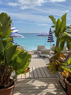 Une terrasse en bois qui mène à une plage agrémentée de parasols à rayures bleues et blanches et de chaises longues.