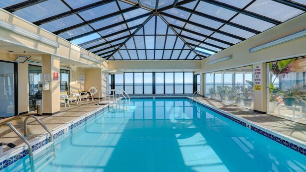 Piscina interna com teto de vidro, vista clara do céu e área ao redor da piscina com cadeiras e aviso proibindo saltos.