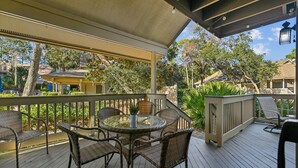 A covered outdoor patio with wicker furniture, a glass table, and a view of a residential area with houses and trees.