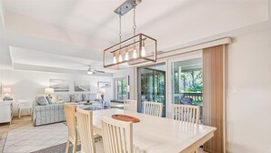 A modern kitchen with a dining area, featuring a large table and chairs, a hanging light fixture, and a view of the outdoors.