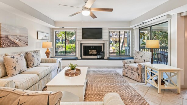 A spacious living room with a fireplace, a ceiling fan, and large windows offering a view of the outdoors.