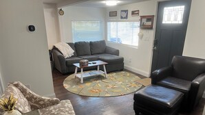 Un salon avec un canapé gris, une table basse, un tapis à motifs floraux et un fauteuil en cuir noir.