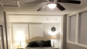 Une chambre à coucher avec un lit, un ventilateur de plafond, une table de chevet avec une lampe, un tapis à motifs floraux et une fenêtre avec des stores.