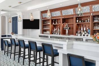 A hotel room with a large bed, a teal sofa, a wooden nightstand, a desk lamp, a chair, a painting of a sailboat, and a large window with a view of a building and greenery. at Embassy Suites by Hilton Charleston Harbor Mt. Pleasant