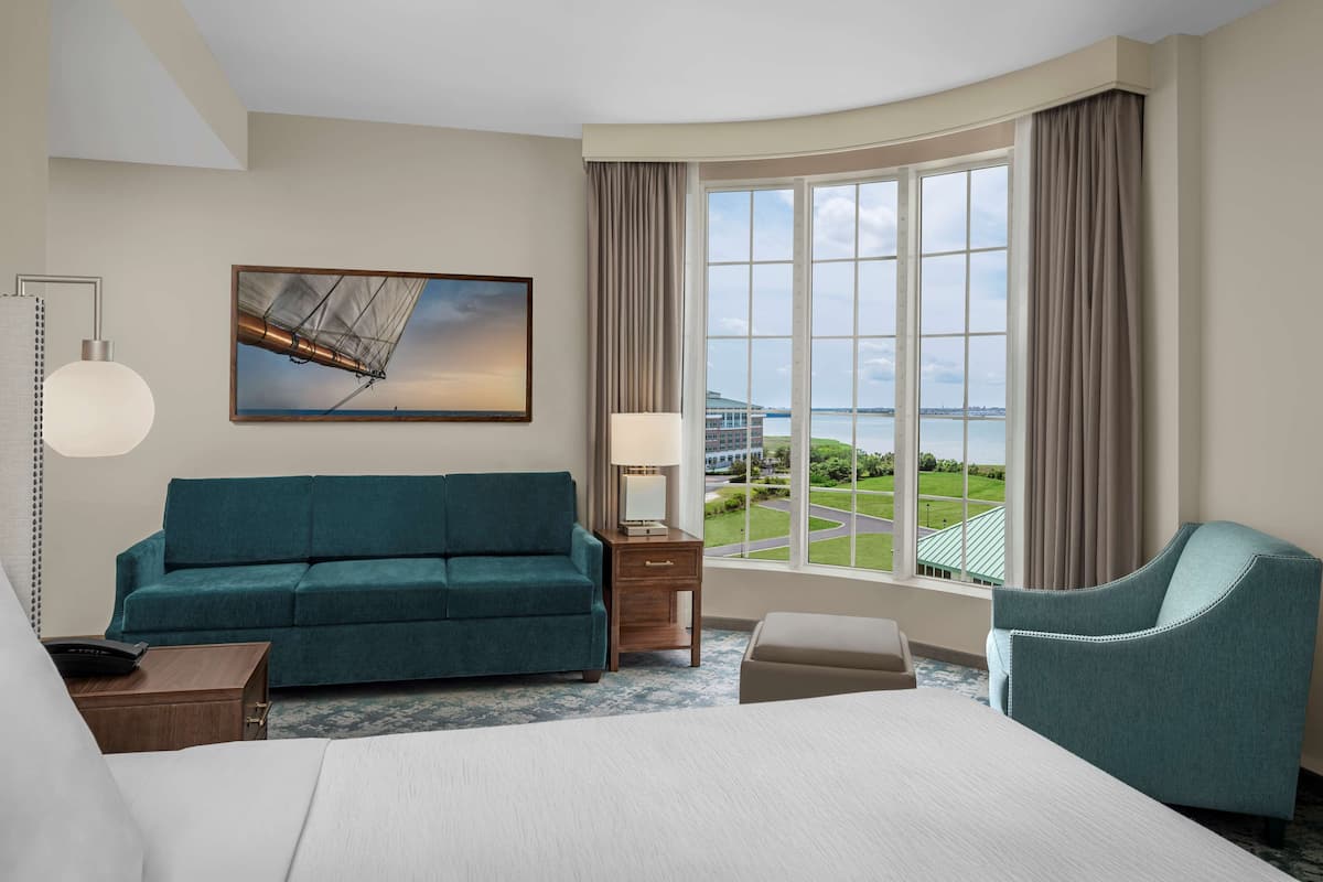 Une chambre d’hôtel avec un grand lit, un canapé de couleur turquoise, une table de chevet en bois, une lampe de bureau, une chaise, un tableau représentant un voilier et une grande fenêtre donnant sur un immeuble et de la verdure.