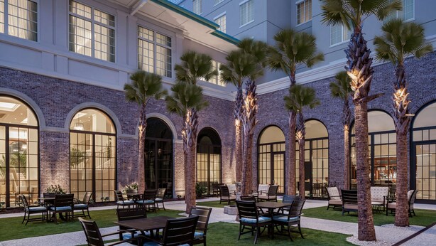 An outdoor patio with tables and chairs, palm trees, and a brick building with large windows. - Embassy Suites by Hilton Charleston Harbor Mt. Pleasant (Mount Pleasant)