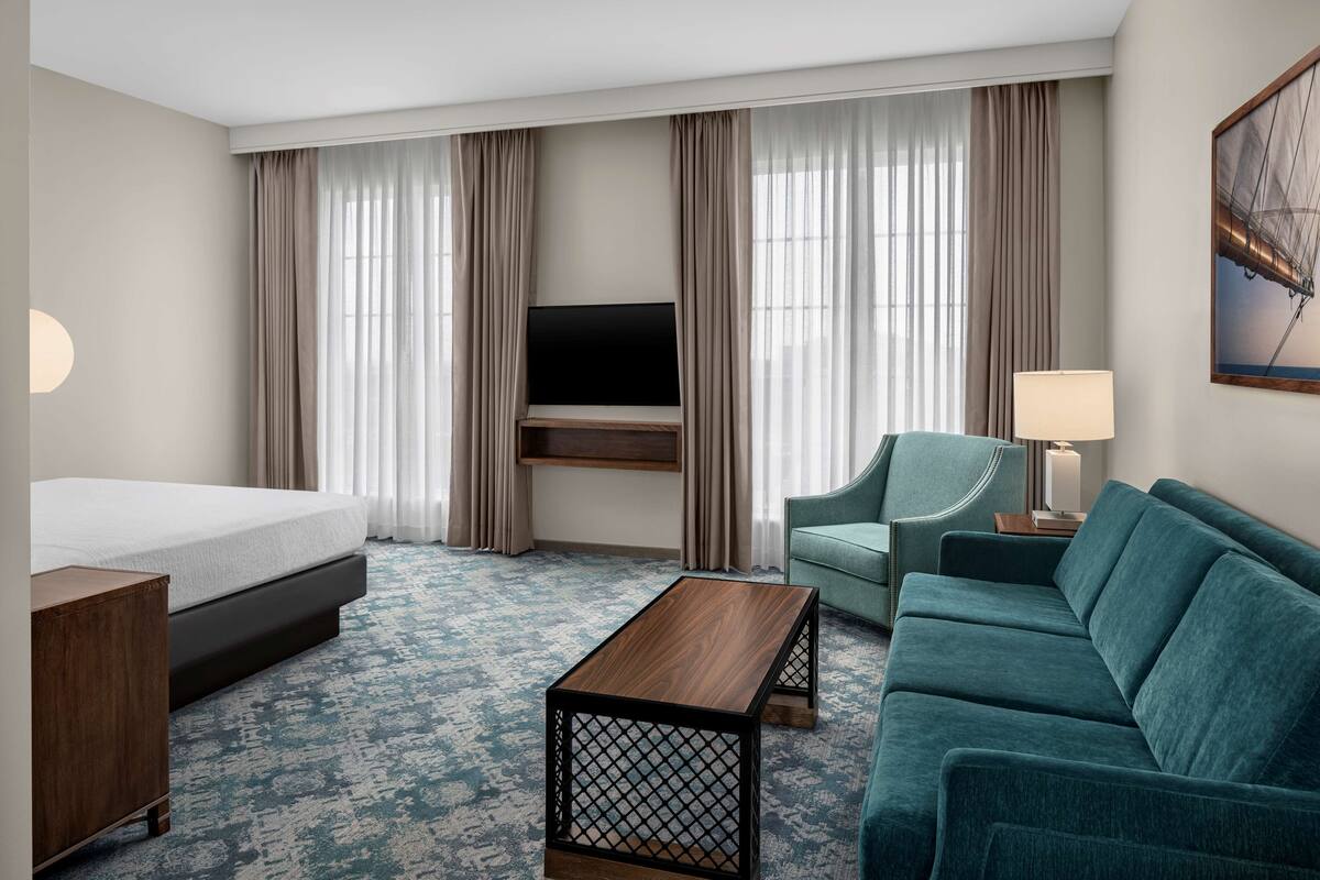 Une chambre d’hôtel comprenant un lit, un canapé, une table basse, un téléviseur fixé au mur et une fenêtre avec des rideaux.