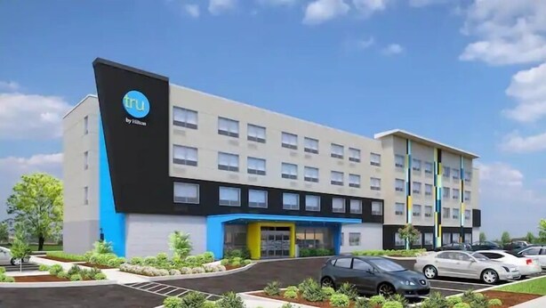A hotel with a modern design, featuring a prominent logo on the facade, a central entrance, and a parking area with several cars.