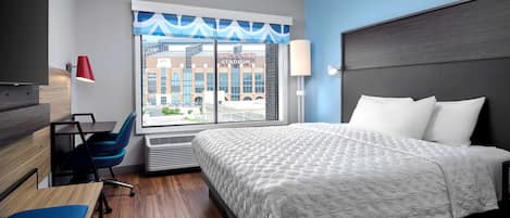Habitación de hotel con una cama grande, un escritorio, una silla, una ventana con vistas a un edificio y una lámpara.