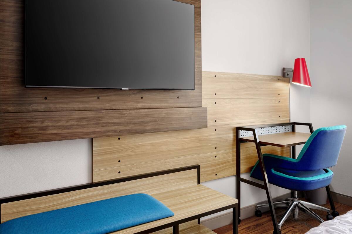 Une chambre d’hôtel moderne équipée d’un téléviseur à écran plat fixé au mur, d’un bureau en bois avec une chaise, d’un banc et d’une lampe de bureau rouge.