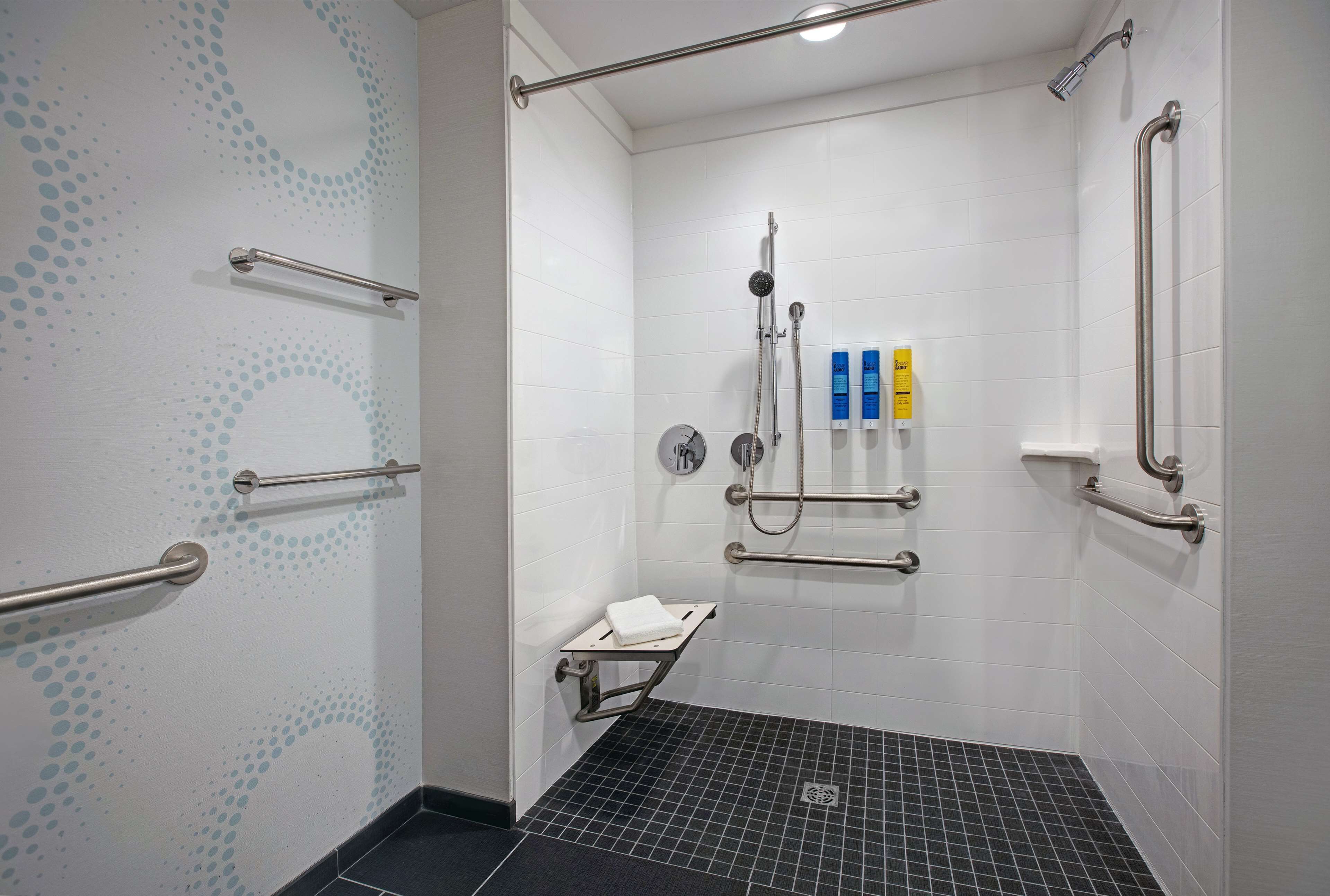 A modern bathroom with white tiled walls, a black tiled floor, and multiple handrails for accessibility.