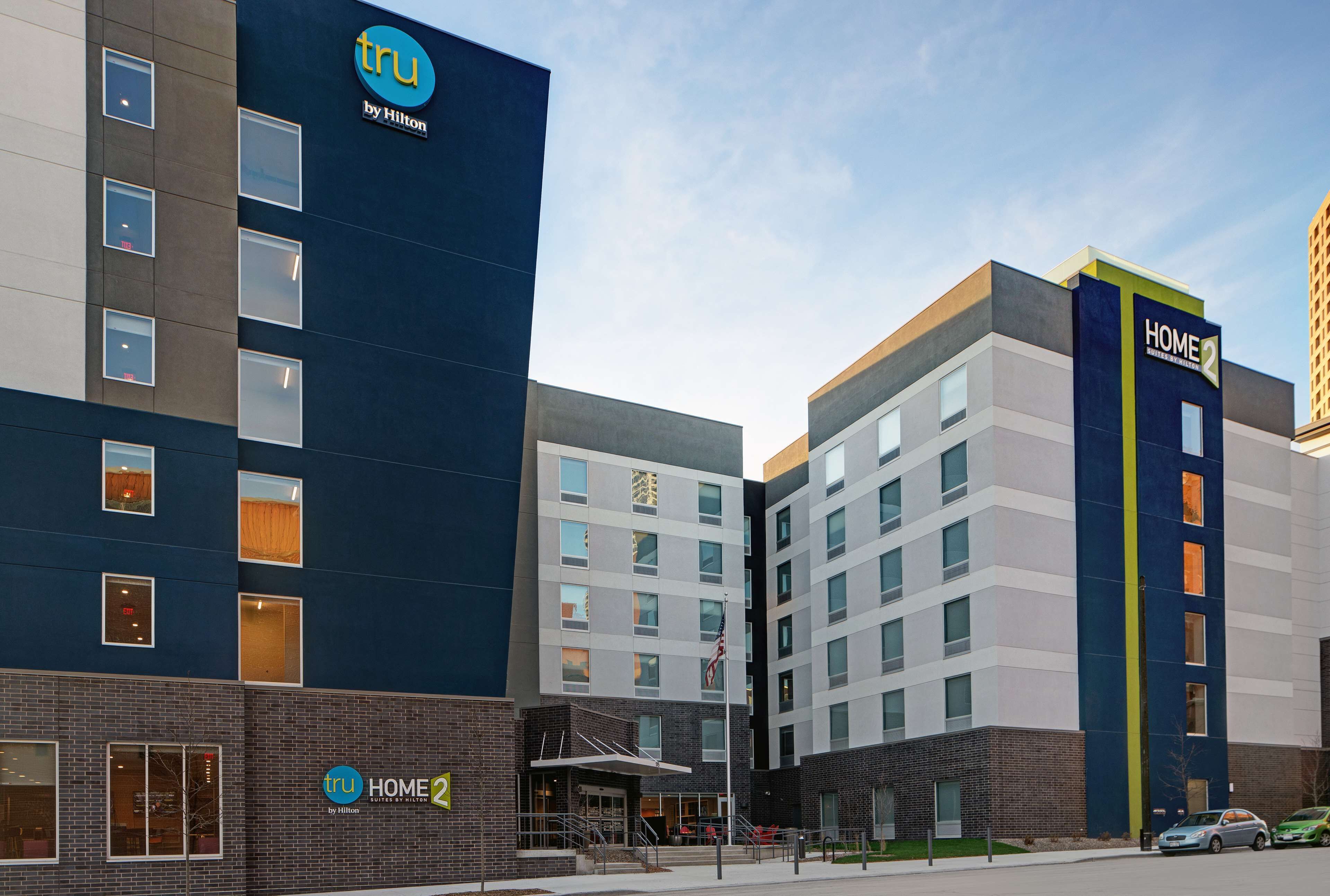 Un hôtel moderne à plusieurs étages, avec une enseigne bien visible « tru by Hilton » et une enseigne « Home 2 » indiquant une section ou une commodité particulière.