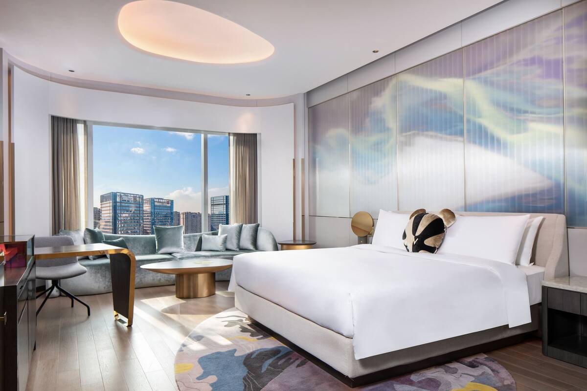A modern hotel room with a large bed, a sofa, a desk, and a view of the cityscape.