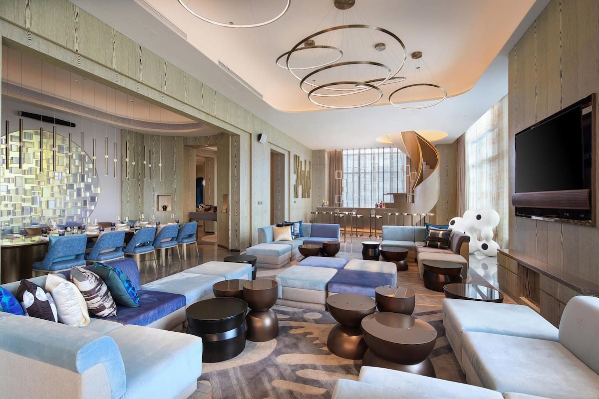 A spacious hotel lobby with a modern design, featuring a large seating area with sofas and armchairs, a bar area, and a large flat-screen TV mounted on the wall.
