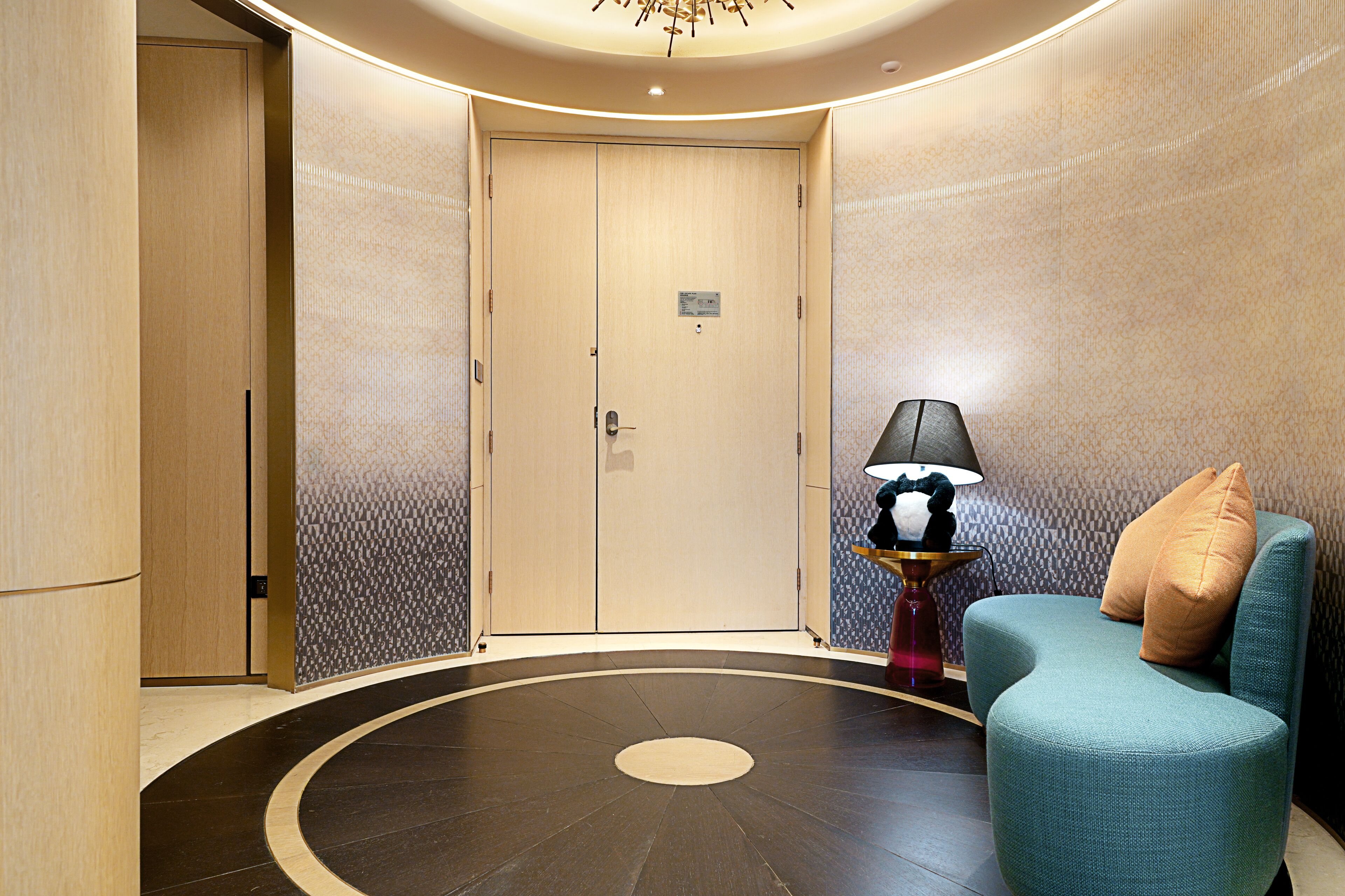 A hotel lobby with a wooden door, a teal armchair with a pillow, a floor lamp, and a patterned wall.