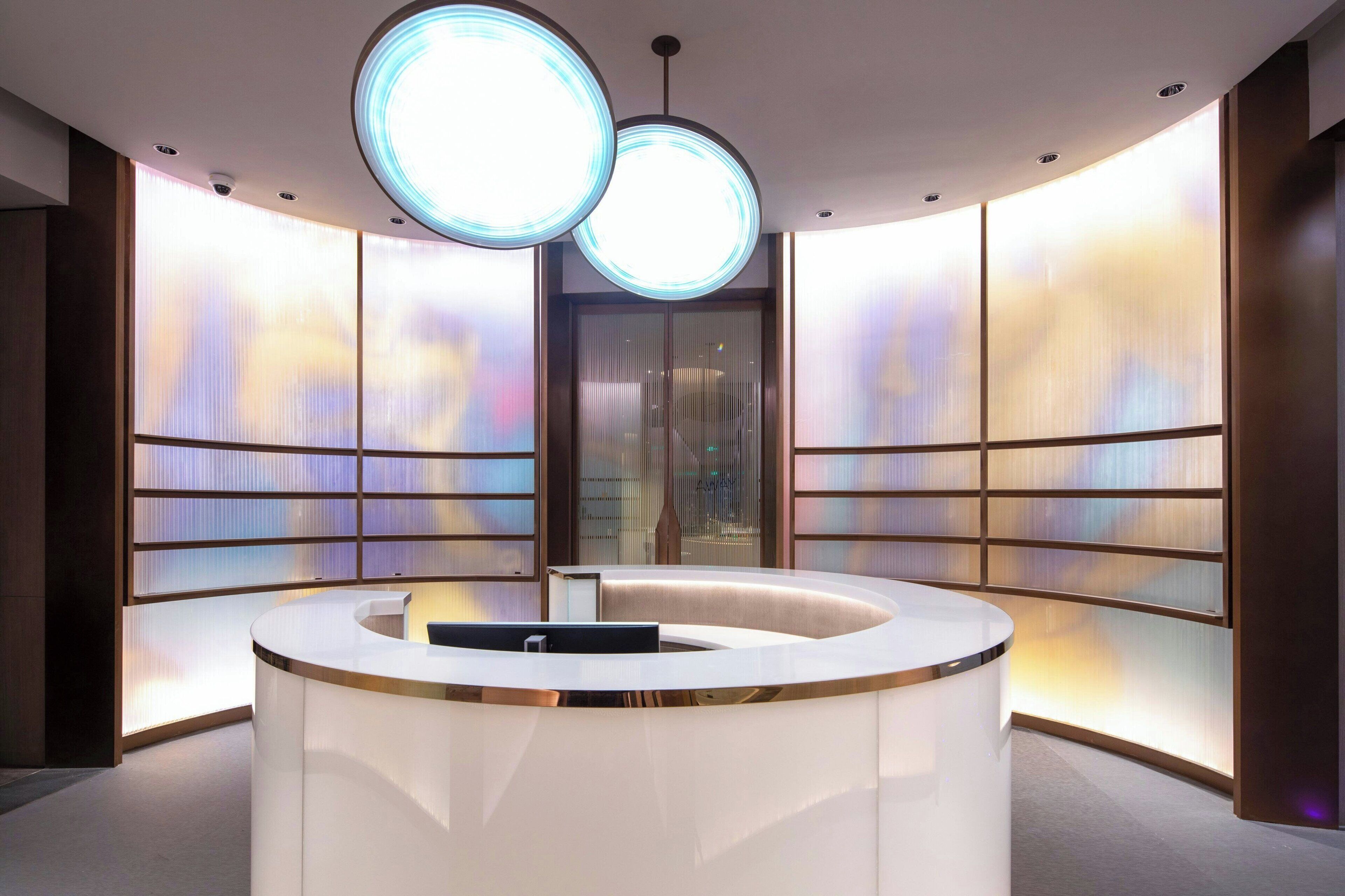 A modern reception area with a curved white reception desk, two large circular ceiling lights, and a view of a building through a large window with frosted glass panels.