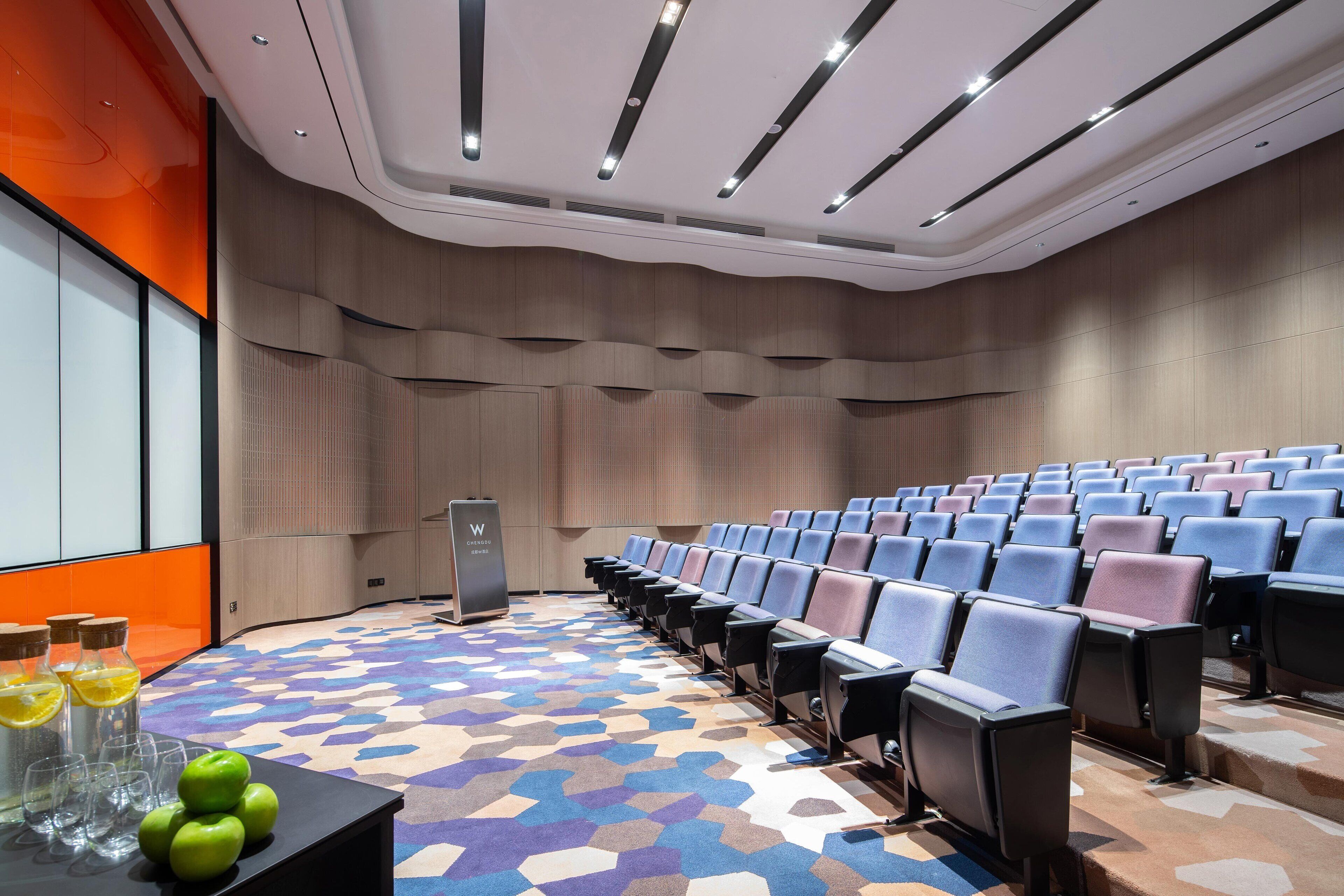 Un salón de conferencias con filas de sillas, un atril y una pantalla para proyecciones.