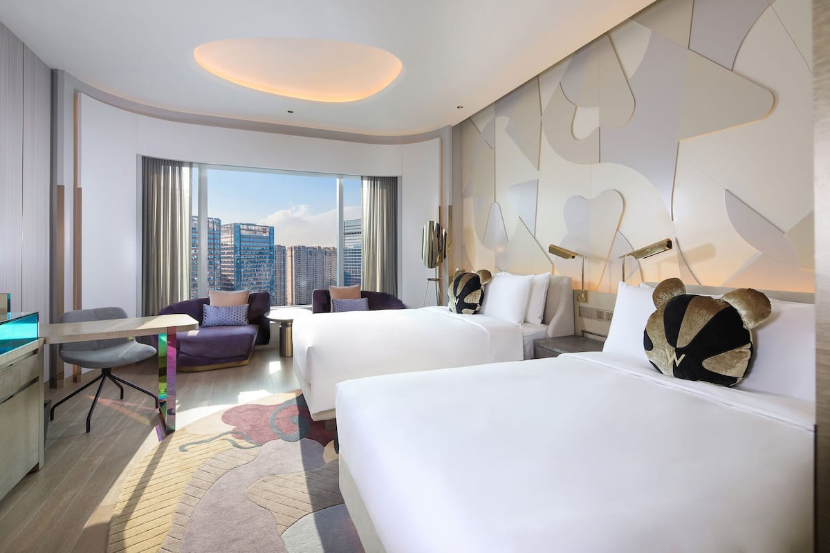 A hotel room with two beds, a desk, a chair, and a view of the cityscape.