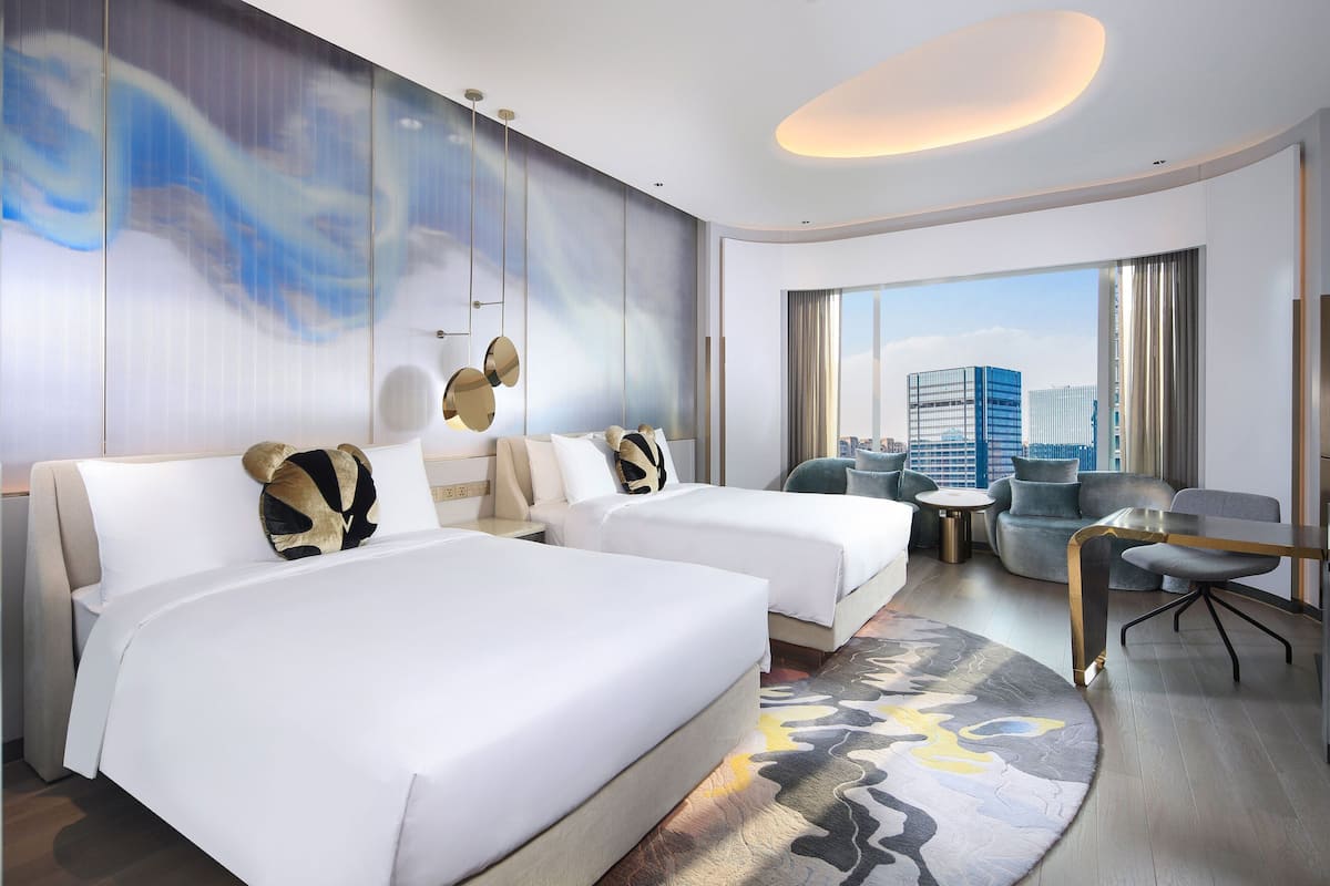 A modern hotel room with two beds, a seating area, and a large window offering a city view.