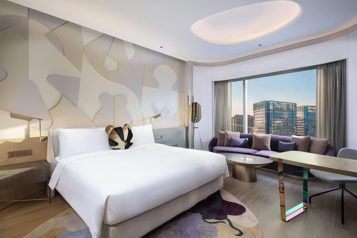 A modern hotel room with a large bed, a sofa, a dining table, and a city view through large windows.