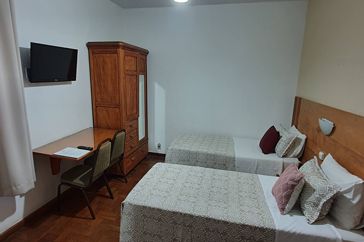 A small hotel room with two single beds, a wooden wardrobe, a desk with a chair, and a TV mounted on the wall.