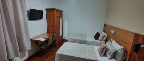 A small hotel room with two single beds, a wooden wardrobe, a desk with a chair, and a TV mounted on the wall.