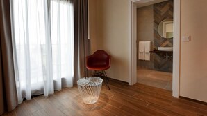 Ein Hotelzimmer mit Holzboden, einem roten Stuhl, einem Glastisch und Blick ins Badezimmer durch eine geöffnete Tür.