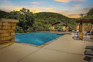 A swimming pool with a stone wall on one side, a covered patio area with outdoor furniture, and a view of a forested hillside. - Miracle Lodge | Pool, Hot Tub & Scenic Views (Leakey)