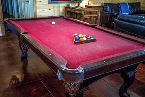 A pool table with a white cue ball and a rack of colored balls in a room with wooden flooring and a leather couch. - Miracle Lodge | Pool, Hot Tub & Scenic Views (Leakey)