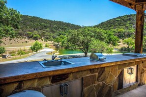 A stone bar with a sink and a grill, overlooking a lake and forested hills. - Miracle Lodge | Pool, Hot Tub & Scenic Views (Leakey)