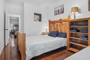 A bedroom with a wooden headboard, a bed with a white quilt, a nightstand with a lamp, and a shelf with folded towels. - Miracle Lodge | Pool, Hot Tub & Scenic Views (Leakey)