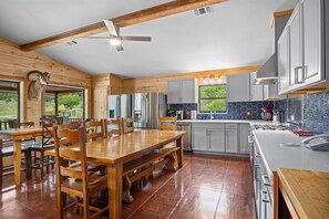 A modern kitchen with wooden cabinetry, a dining area with a wooden table and chairs, and a ceiling fan. - Miracle Lodge | Pool, Hot Tub & Scenic Views (Leakey)