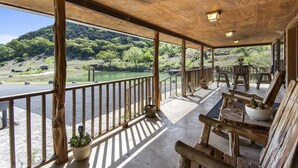 Terraza de madera con vistas a un río y colinas. Se observan sillas y mesas de madera, además de una barandilla a lo largo del borde.