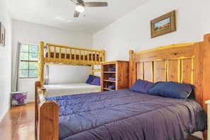 A room with a bunk bed, a single bed, wooden headboard, a shelf unit, and a window with curtains. - Miracle Lodge | Pool, Hot Tub & Scenic Views (Leakey)