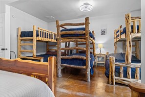 A room with bunk beds made of wood, a wooden chair, a bedside table with a lamp, and a bed with a white quilt. - Miracle Lodge | Pool, Hot Tub & Scenic Views (Leakey)