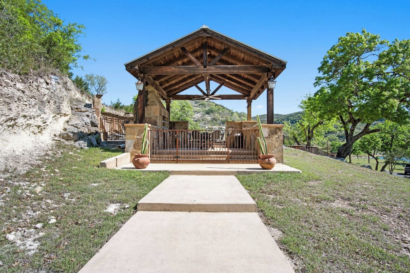 A wooden gazebo with a concrete pathway leading to it, surrounded by grass and trees.
