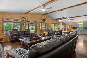 A spacious living room with a large leather sofa, a wooden coffee table, and a fireplace. There are multiple windows providing natural light and views of the outdoors. - Miracle Lodge | Pool, Hot Tub & Scenic Views (Leakey)