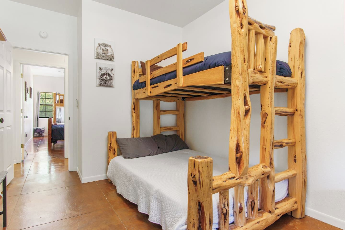 A bunk bed with a wooden frame and a single bed below. There is a door leading to another room and a window with curtains.