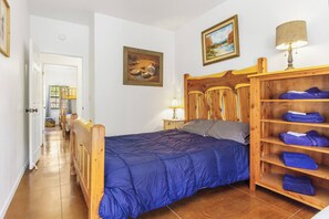 A bedroom with a wooden bed, a nightstand with a lamp, a shelf with folded towels, and two paintings on the wall. - Miracle Lodge | Pool, Hot Tub & Scenic Views (Leakey)