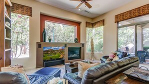 A living room with a fireplace, leather sofas, a television, and large windows with views of trees.